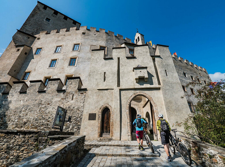 Biken Schloss Bruck Zwei Biker schieben ihre Räder durch einen steinernen Bogen am Eingang des Schloss Bruck in Lienz.