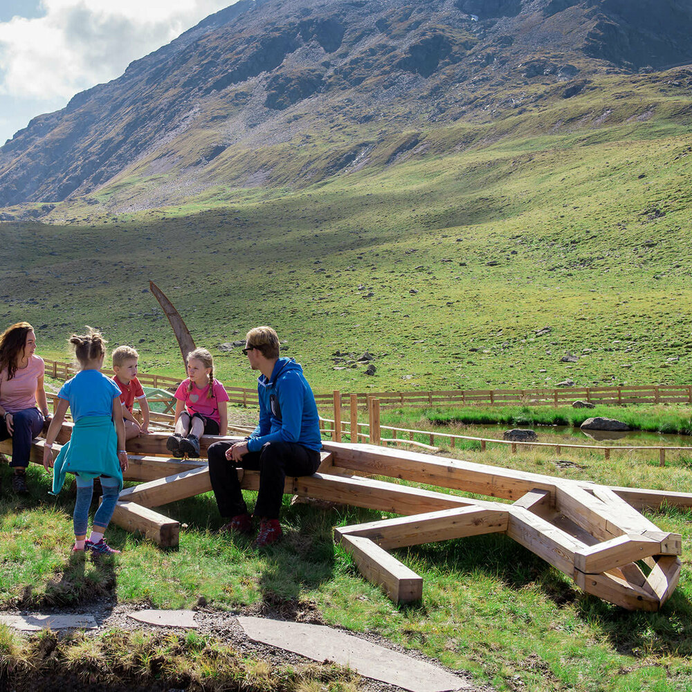 Wassermythos Ochsenlacke St. Jakob Brunnalm Eine Familie sitzt bei der Spielarena des Wassermythos Ochsenlacke im Skizentrum St. Jakob i. D.. Die Sonne lässt die Umgebung und die Berge im Hintergrund in einem besonderes warmen Licht erstrahlen.