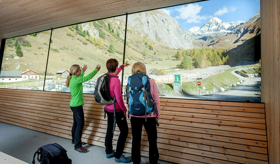 Nationalpark Glocknerpanorama „Am Parkplatz Glocknerwinkel in Kals stehen drei Frauen und schauen durch eine Glaswand auf den Großglockner. Die herbstliche Landschaft zeigt bunt gefärbte Bäume.“