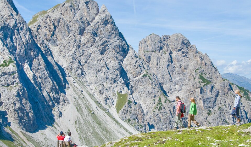 Mehrere Personen wandern in den Lienzer Dolomiten mit beeindruckender Bergkulisse. Zwei Personen sitzen auf einer Holzbank und blicken auf eine steile Felswand.