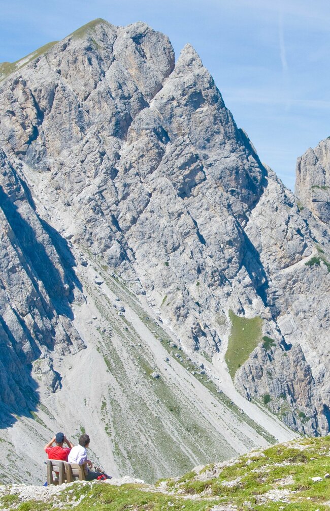 Mehrere Personen wandern in den Lienzer Dolomiten mit beeindruckender Bergkulisse. Zwei Personen sitzen auf einer Holzbank und blicken auf eine steile Felswand.