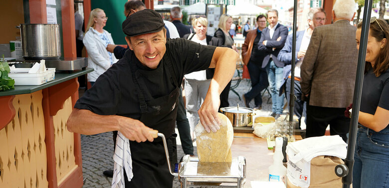 Koch Ernst Muhr dreht auf seinem Stand den Nudelteig durch die Nudelmaschine. Einige Menschen sehen ihm dabei zu.