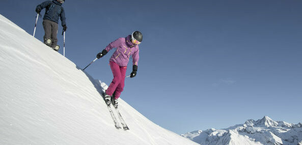 Zwei Skifahrer:innen stürzen sich im freien Gelände in einen steilen Hang mit Pulverschnee.