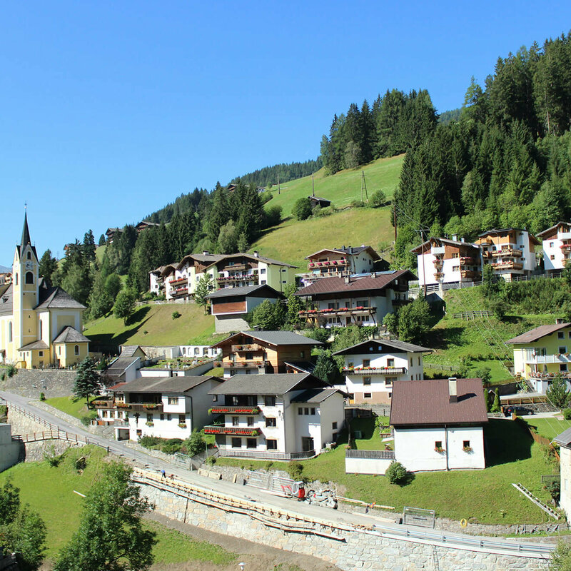 Das Bild zeigt das Dorf Außervillgraten mit seiner Kirche und einer Wohnsiedlung auf saftig grünem Untergrund, gelegen unter strahlend blauem Himmel.