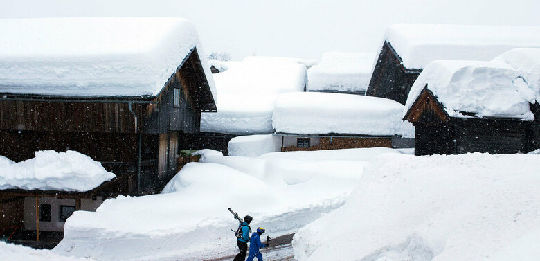 Dicke Schneehauben auf den Dächern im winterlichen Obertilliach. Zwei Skifahrer gehen zu Fuß über geräumte aber von meterhohen Schneewänden gesäumte Dorfgasse.