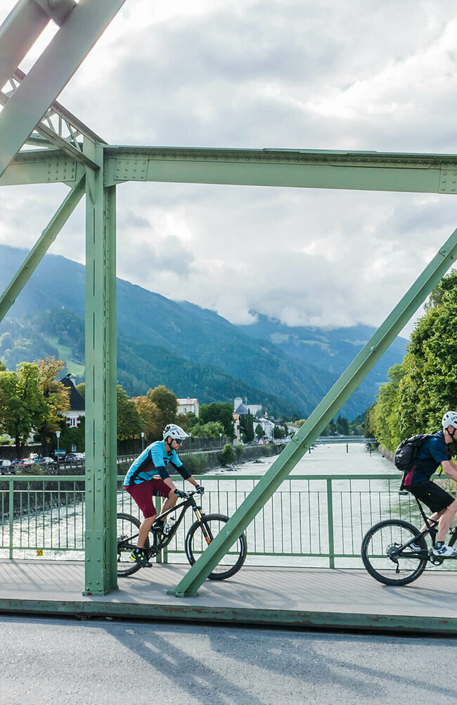 Zwei Radfahrer fahren über eine asphaltierte Brücke über der Isel in der Stadt Lienz.