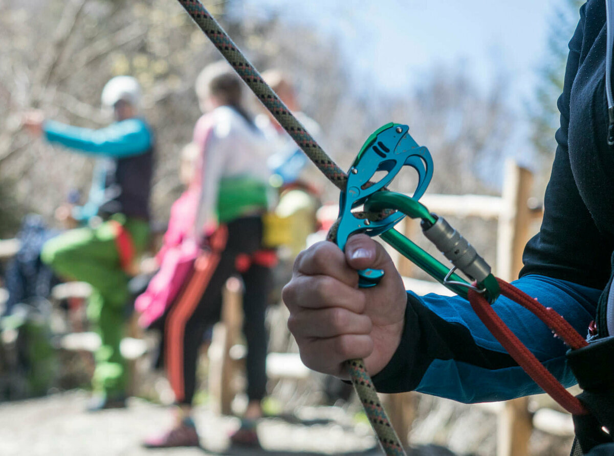 Karabiner Nahaufnahme von einem Kletter-Sicherungsgerät, einem Seil und bunten Karabinern. Im Hintergrund stehen mehrere Personen.