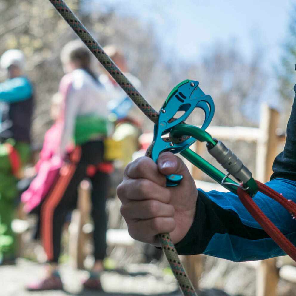Nahaufnahme von einem Kletter-Sicherungsgerät, einem Seil und bunten Karabinern. Im Hintergrund stehen mehrere Personen.
