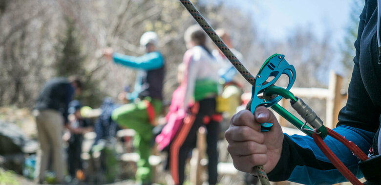Karabiner Nahaufnahme von einem Kletter-Sicherungsgerät, einem Seil und bunten Karabinern. Im Hintergrund stehen mehrere Personen.