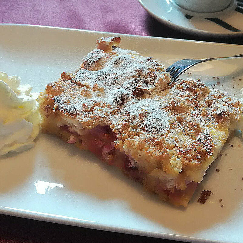 Ein Stück Streuselkuchen auf einem weißen Teller mit zwei Portionen Sahne daneben.