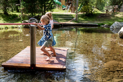 Spielende Kinder im Teich des Wassernaturspielplatzes St. Jakob i. D.. Sie ziehen sich auf einem kleinen Holzfloss anhand eines gespannten Seiles über das Wasser. Die Sonne lässt den Teich und die Umgebung in einem warmen Licht erstrahlen.