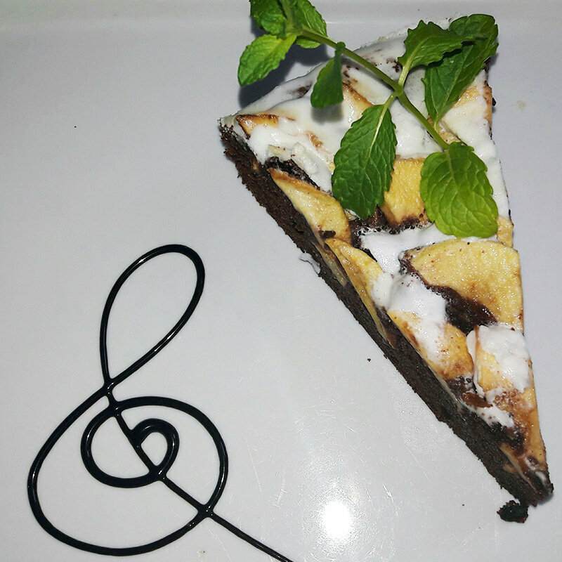 Ein Stück Schoko-Apfelkuchen auf einem Teller mit einem Notenschlüssel aus Schoko-Soße verziert.