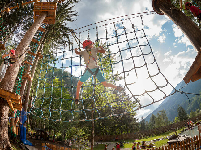 Kinder an einer Kletterstation im Hochseilgarten der Galitzenklamm bei sonnigem Wetter. Im Hintergrund spielen mehrere Personen am Wasserspielplatz. 