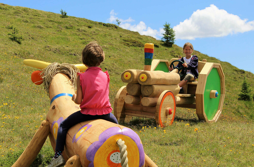 Zwei Mädchen, die auf einer bunten Holzkuh bzw. auf einem bunten Holztraktor sitzen. Dahinter ein grasbewachsener Berghang mit spärlichem Bewuchs und einem leicht bewölkten Himmel.
