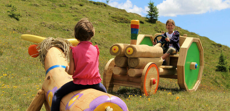 Kindermeile Golzentipp Obertilliach Zwei Mädchen, die auf einer bunten Holzkuh bzw. auf einem bunten Holztraktor sitzen. Dahinter ein grasbewachsener Berghang mit spärlichem Bewuchs und einem leicht bewölkten Himmel.