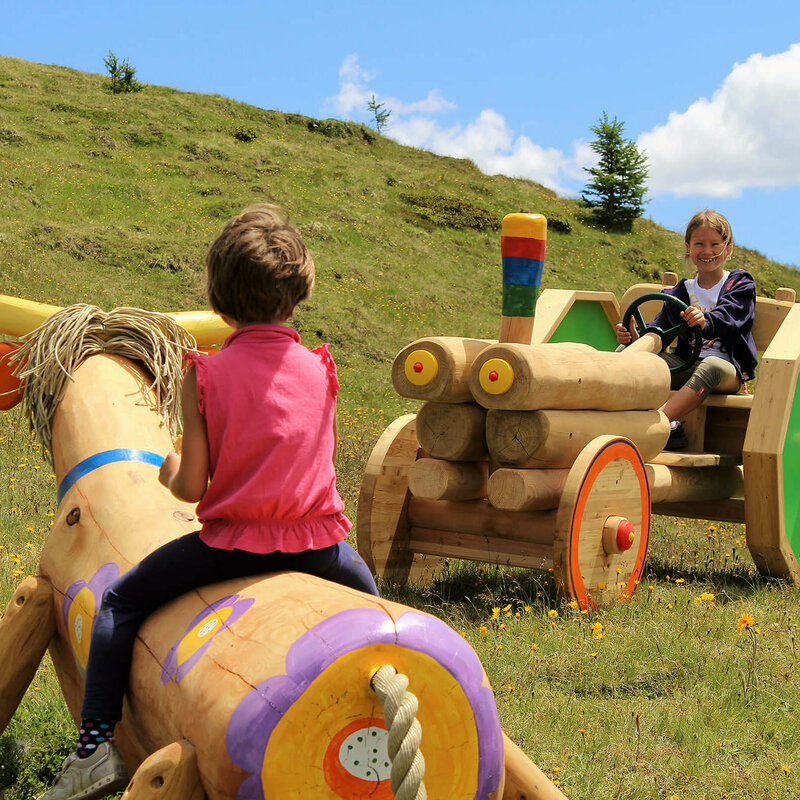 Zwei Mädchen, die auf einer bunten Holzkuh bzw. auf einem bunten Holztraktor sitzen. Dahinter ein grasbewachsener Berghang mit spärlichem Bewuchs und einem leicht bewölkten Himmel.