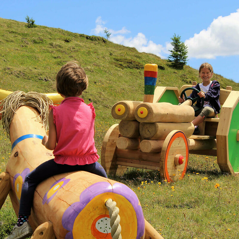 Kindermeile Golzentipp Obertilliach Zwei Mädchen, die auf einer bunten Holzkuh bzw. auf einem bunten Holztraktor sitzen. Dahinter ein grasbewachsener Berghang mit spärlichem Bewuchs und einem leicht bewölkten Himmel.
