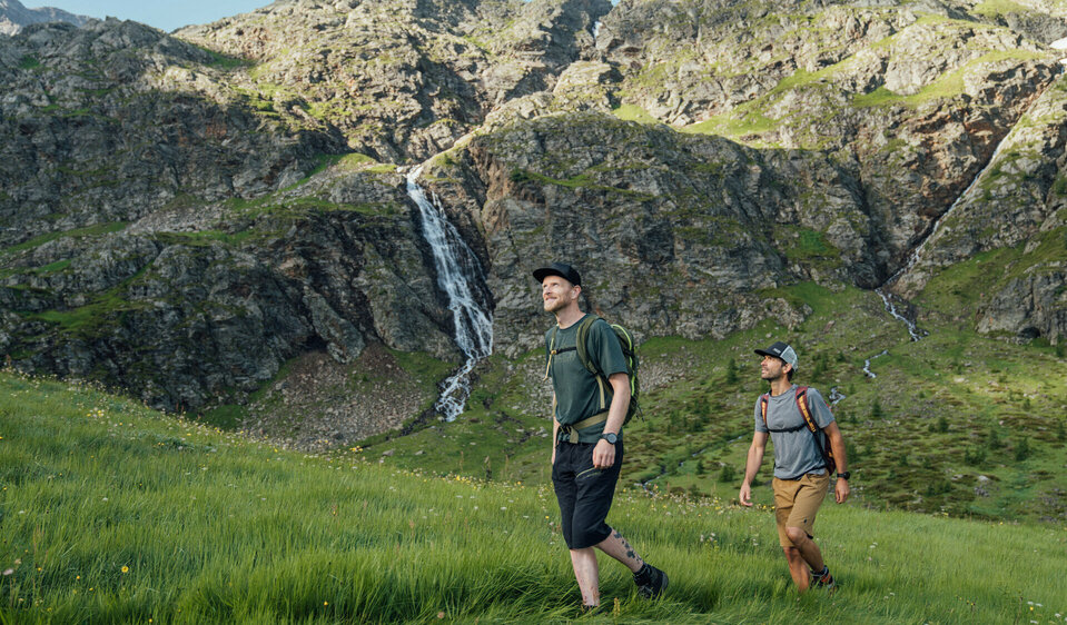 Zwei Männer wandern in Osttirols Bergen hinauf zum Geigensee durch hohes Bergwiesengras. Im Hintergrund ist ein kleiner Wasserfall zu sehen, der talwärts rinnt.