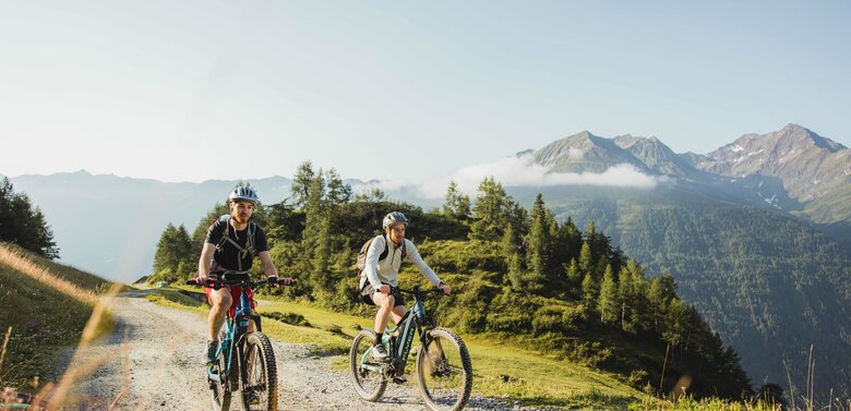 Biketour Timmeltal Praegraten Zwei junge E-Biker auf blau-grauen Bikes die bergwärts auf einem leicht ansteigenden Schotterweg fahren. Die Sonne scheint ihnen in den Rücken. Den Hintergrund bildet eine kleine, leicht bewaldete Erhebung hinter dieser es tief ins Tal zu gehen scheint. Ganz im Hinergrund erheben sich zwei Berge, deren Gipfelbereich eine dünne Nebelbank ziert.