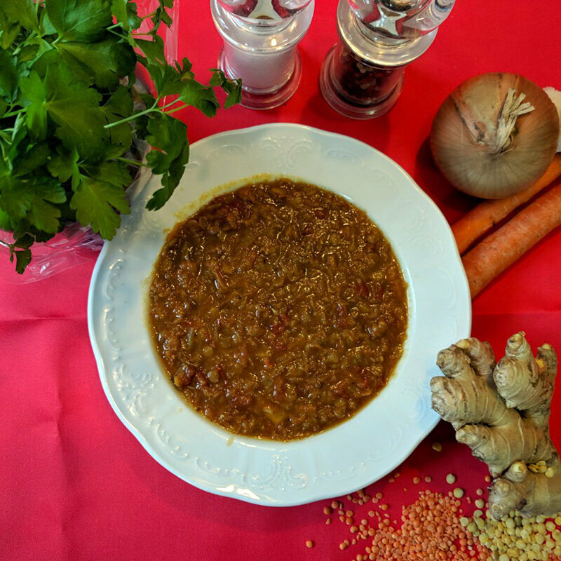 Ein Teller Rote Linsen Suppe mit mehreren Zutaten, wie Ingwer, Zwiebel, Knoblauch, Karotte, sowie Salz und Pfeffer daneben. 