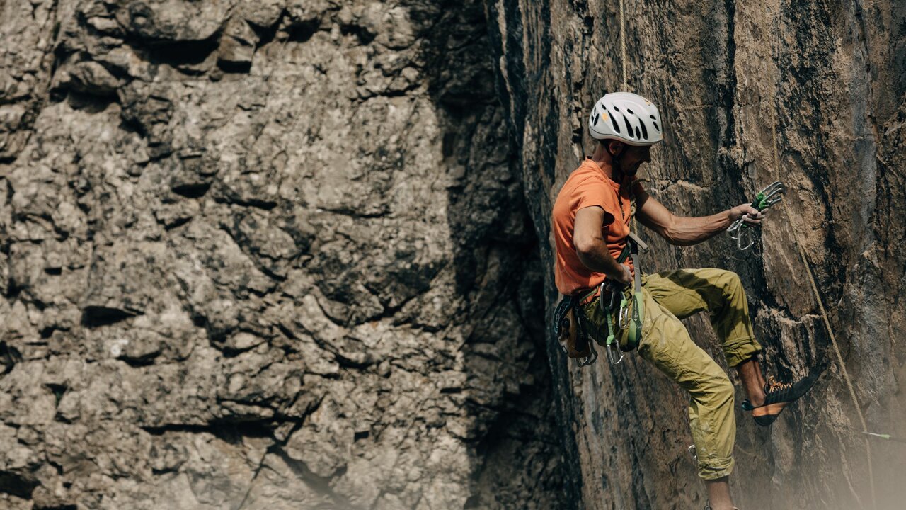 Ein Kletterer mit orangenem Shirt und weißem Helm, hängt an einer steilen Felswand am Laserz Klettersteig.
