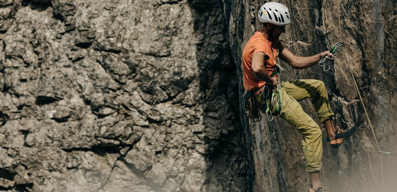 Ein Kletterer mit orangenem Shirt und weißem Helm, hängt an einer steilen Felswand am Laserz Klettersteig.