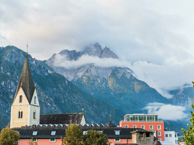 Blick auf die Hausdächer und den Kirchturm in Lienz mit beeindruckender Bergkulisse im Hintergrund.