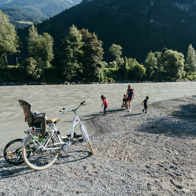Radfahren Eine Familie spielt am Ufer der Isel, im Vordergrund stehen zwei Räder eines in weiß und ein Kinderrad in giftgrün