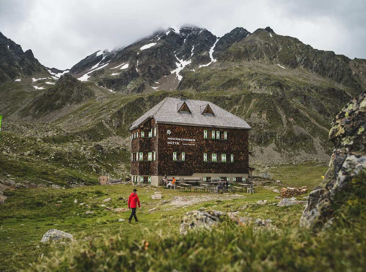 Eine Person mit roter Jacke geht kurz vor der Hochschober Hütte. Links steht eine grüne Alpenvereins-Flagge.