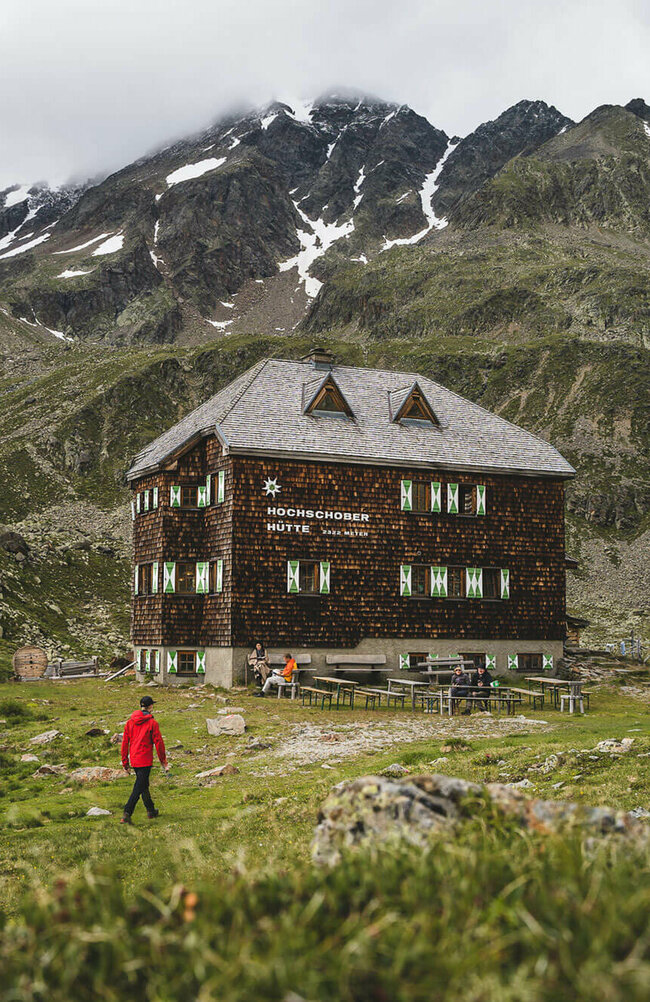 Hochschober Hütte Eine Person mit roter Jacke geht kurz vor der Hochschober Hütte. Links steht eine grüne Alpenvereins-Flagge.