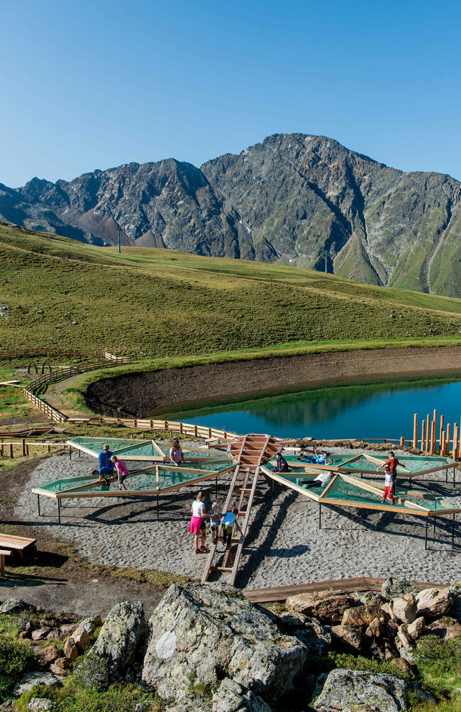 Zu sehen sind mehrere Kinder die auf einem Spielgerät, welches einer überdimensional-großen Libelle ähnlich sieht, beim Wassermythos Ochsenlacke im Skizentrum St. Jakob i. D. spielen. Das sonnige Wetter lässt den sichtbaren Teil des Ufers und die dahinterliegende Bergwelt in einem warmen Licht erstrahlen.