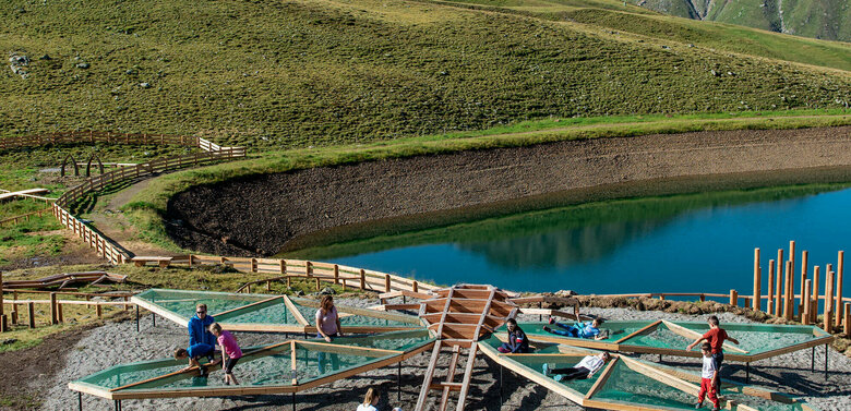 Wassermythos Ochsenlacke St. Jakob Brunnalm Zu sehen sind mehrere Kinder die auf einem Spielgerät, welches einer überdimensional-großen Libelle ähnlich sieht, beim Wassermythos Ochsenlacke im Skizentrum St. Jakob i. D. spielen. Das sonnige Wetter lässt den sichtbaren Teil des Ufers und die dahinterliegende Bergwelt in einem warmen Licht erstrahlen.