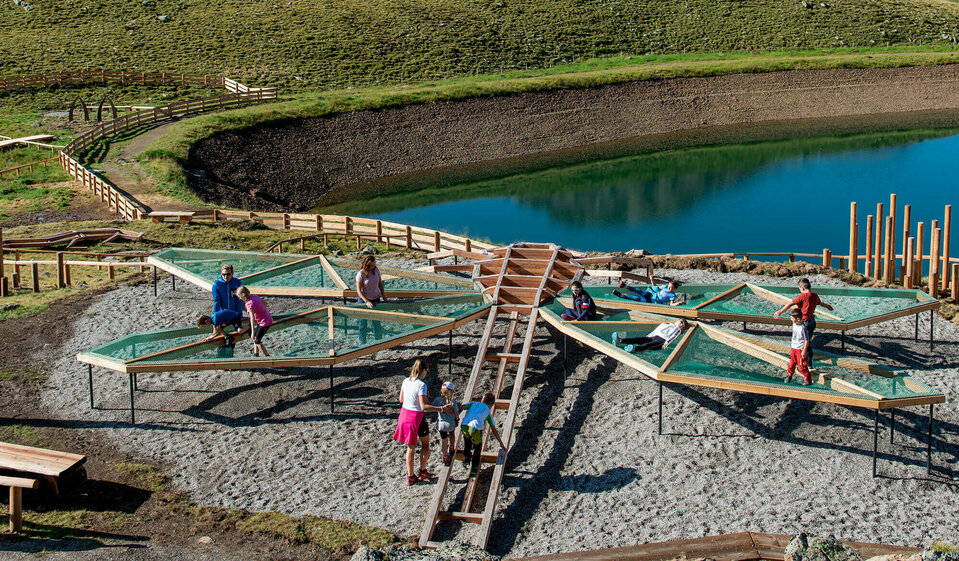 Zu sehen sind mehrere Kinder die auf einem Spielgerät, welches einer überdimensional-großen Libelle ähnlich sieht, beim Wassermythos Ochsenlacke im Skizentrum St. Jakob i. D. spielen. Das sonnige Wetter lässt den sichtbaren Teil des Ufers und die dahinterliegende Bergwelt in einem warmen Licht erstrahlen.