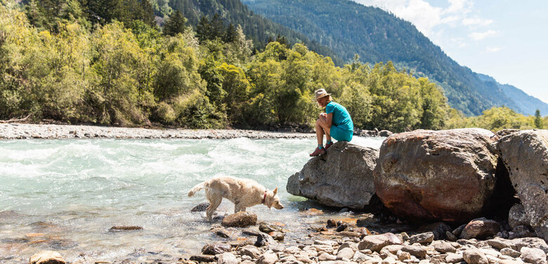 Frau sitzt auf einem großen Stein und schaut in die vorbeifließende Isel, daneben schüttelt ihr Hund das Wasser von sich.