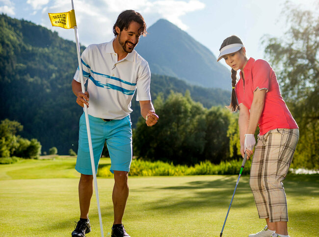 Eine Frau mit Golfschläger locht den Golfball ein im Dolomitengolf Resort. Ein Mann steht nebendran, hält die Fahne und feuert sie an.