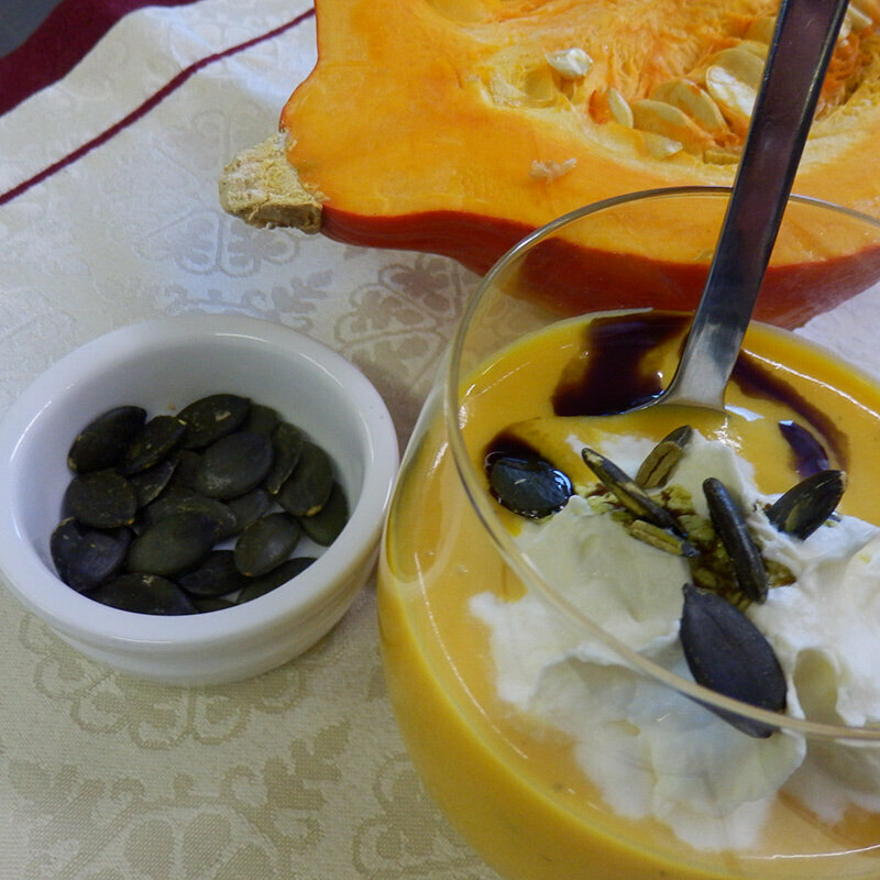 Ein Glas mit Kürbis-Apfel-Suppe gefüllt und Schlag und Kürbiskernen drauf. Daneben eine Schale mit Kürbiskernen und ein halber Kürbis.