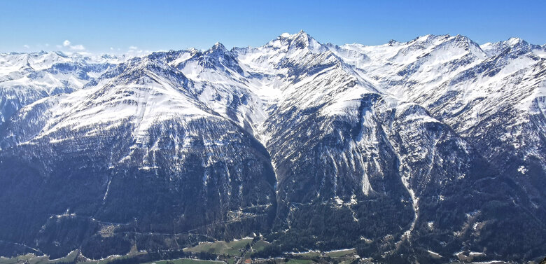 Blick vom vorderen Sajatkopf auf die Gemeinde Prägraten mit hohen Bergen im Hintergrund. Im Tal sind die Wiesen bereits grün, während auf die Berggipfel noch schneebedeckt sind.