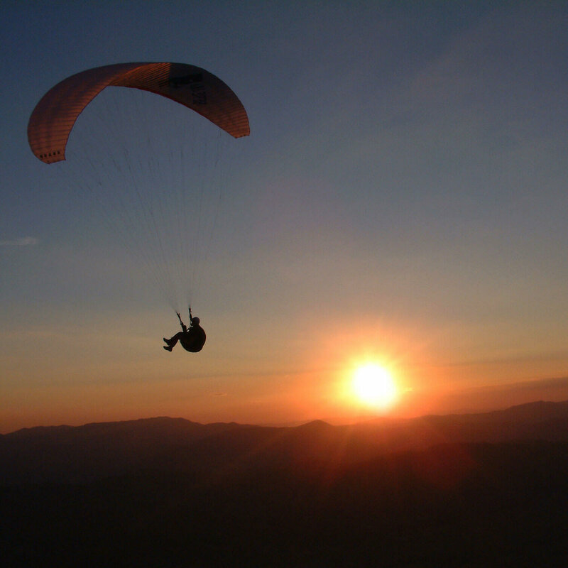 Paragleiter am abendlichen Himmel, dahinter Sonne, die bald am Horizont verschwindet.