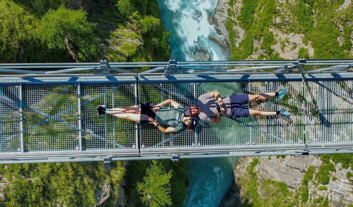 Die Iseltrail-Hängebrücke aus der Vogelperspektive mit zwei Menschen, die darauf liegen und der blauen Isel tief darunter.