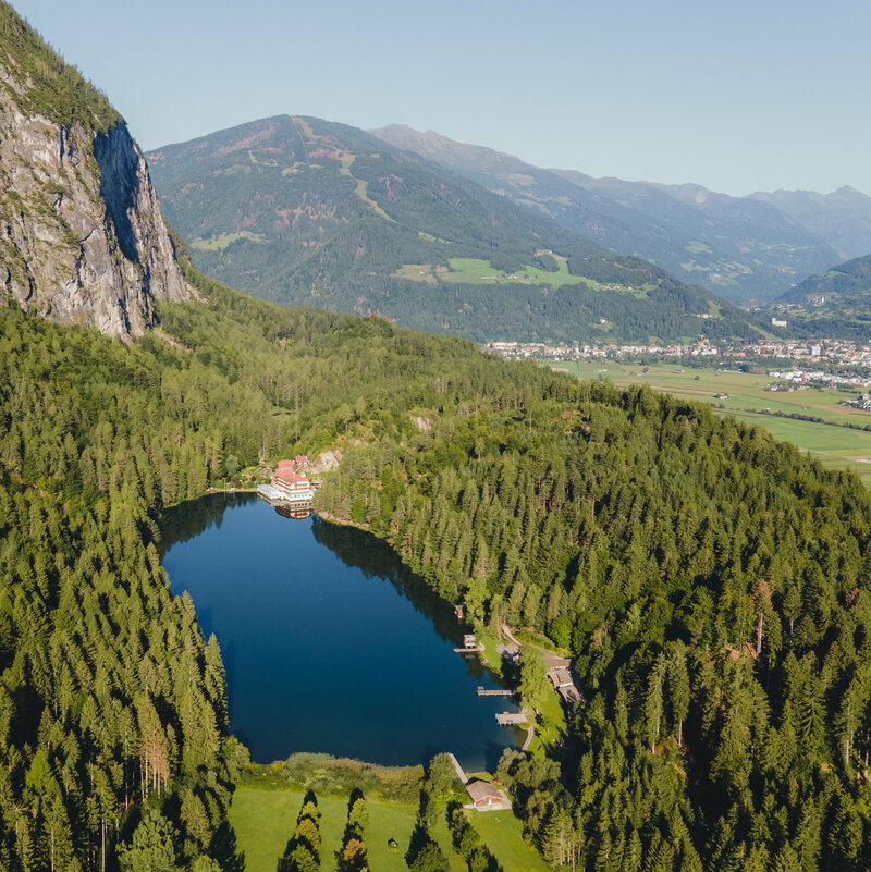 Tristacher See Luftaufnahme vom sanft in den Wald eingebetteten Tristacher See an einem sonnigen Tag, mit Blick auf die Stadt im Hintergrund.