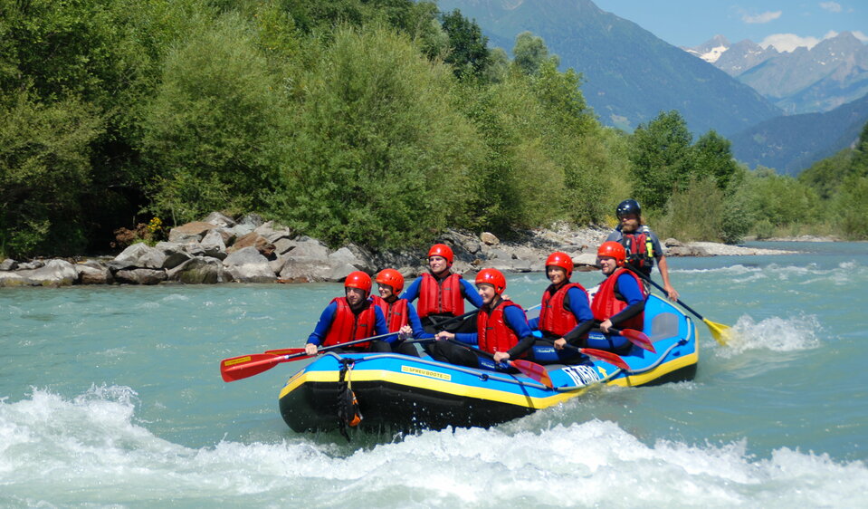Ein Raftingboot mit mehreren Personen in Neoprenanzügen mit roten Schwimmwesten und roten Helmen auf der Isel.