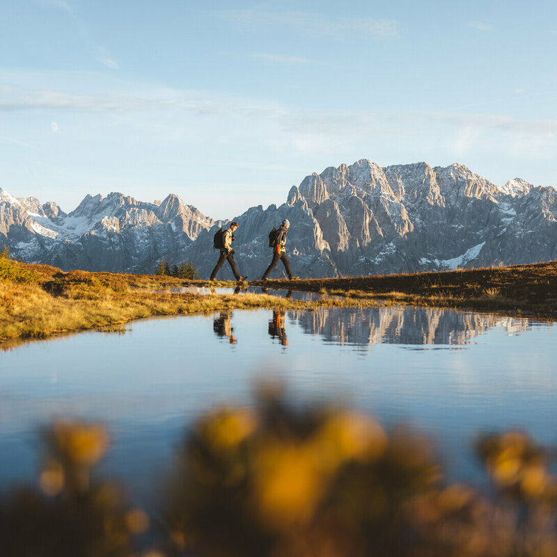 Zwei Personen wandern mit Rucksäcken entlang eines klaren Bergsees. Im Hintergrund erhebt sich eine beeindruckende Alpenkulisse mit schneebedeckten Gipfeln. Das warme Licht der tiefstehenden Sonne beleuchtet die herbstliche Landschaft.