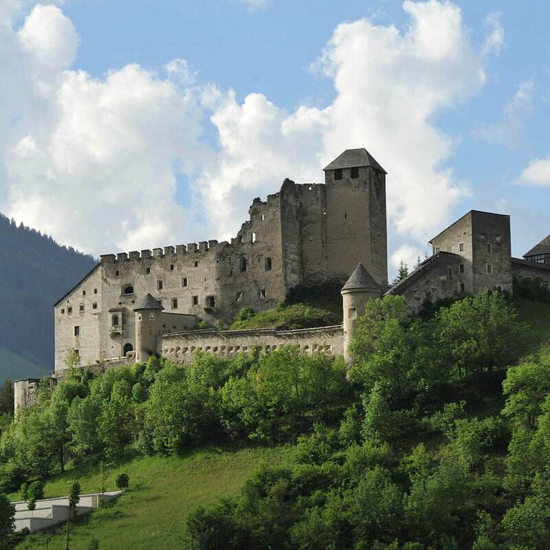 Majestätischer Sommerblick aus dem Tal auf die Burg Heinfels, eingebettet in grüne Laubbäume im satten Sommerkleid