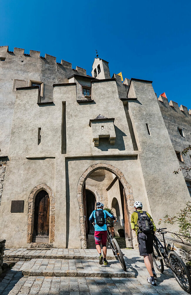Zwei Radfahrer schieben ihre Fahrräder durch einen steinernen Eingangsbogen in das Schloss Bruck in Lienz.
