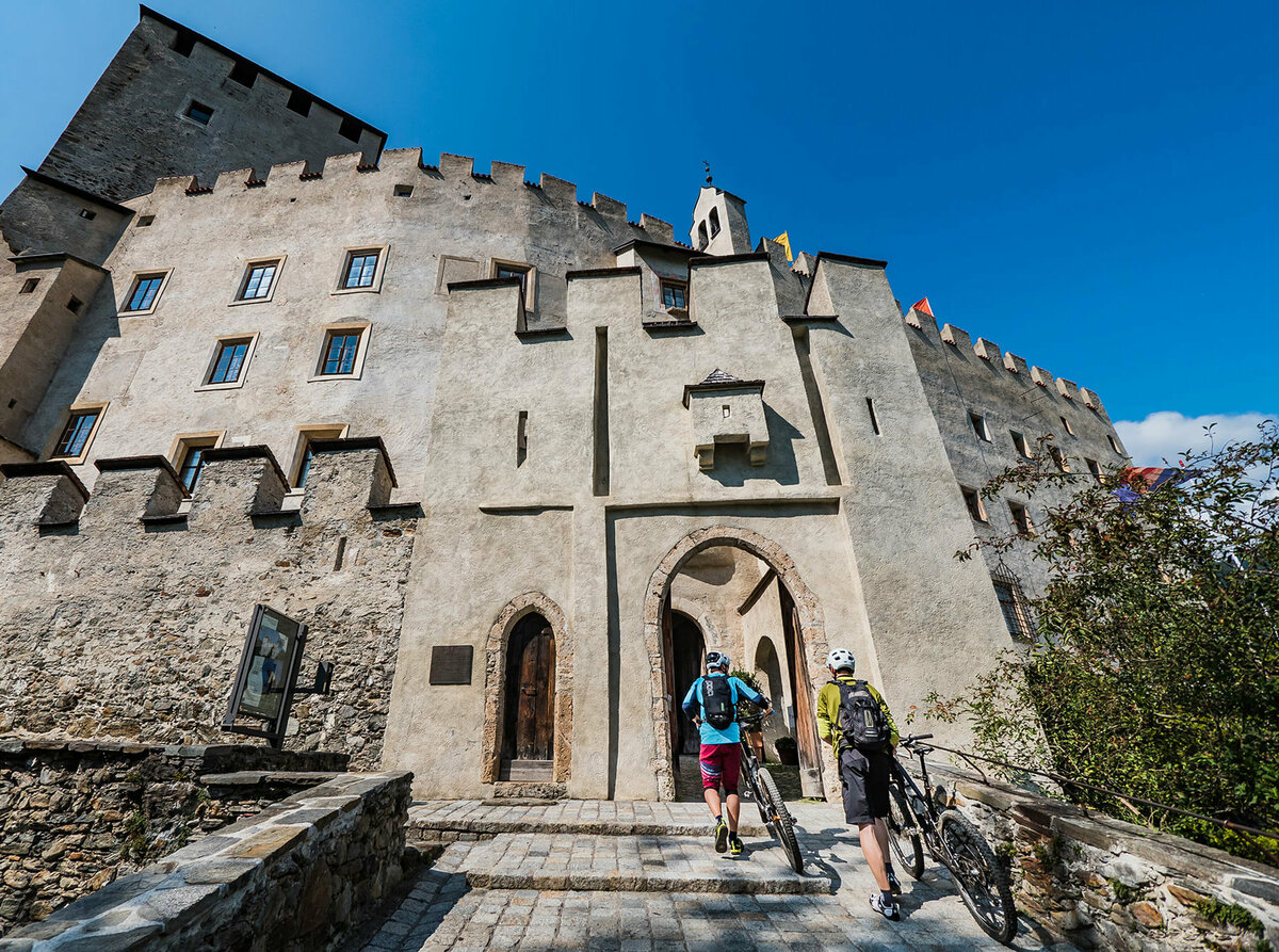 Schloss Bruck Zwei Radfahrer schieben ihre Fahrräder durch einen steinernen Eingangsbogen in das Schloss Bruck in Lienz.