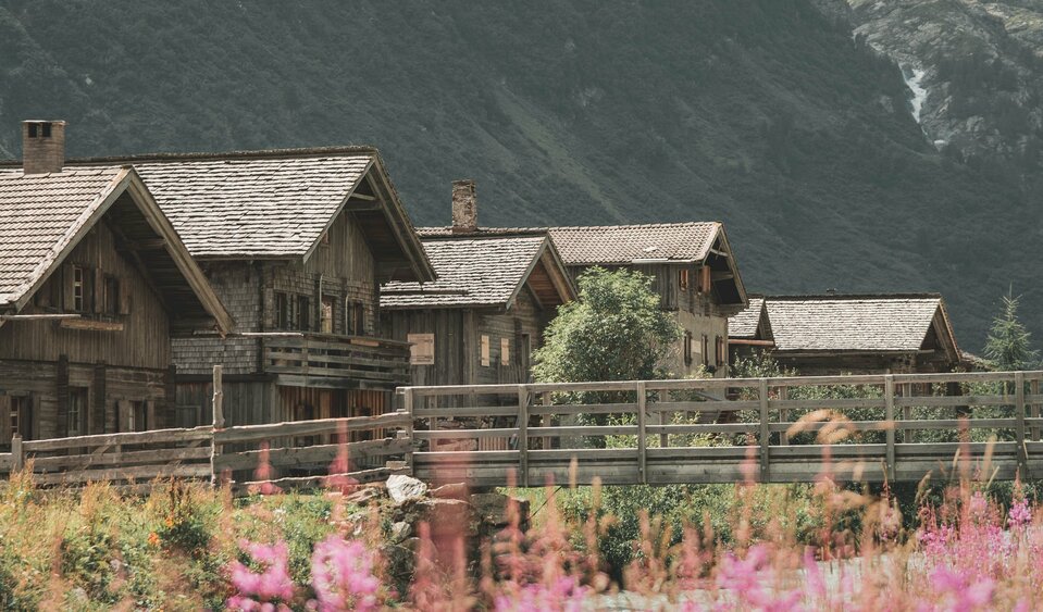 Mehrere alte Holzgebäude vor hohen, schneebedeckten Berggipfeln. Im Vordergrund wachsen rosafarbene Blumen neben einem Fluss.