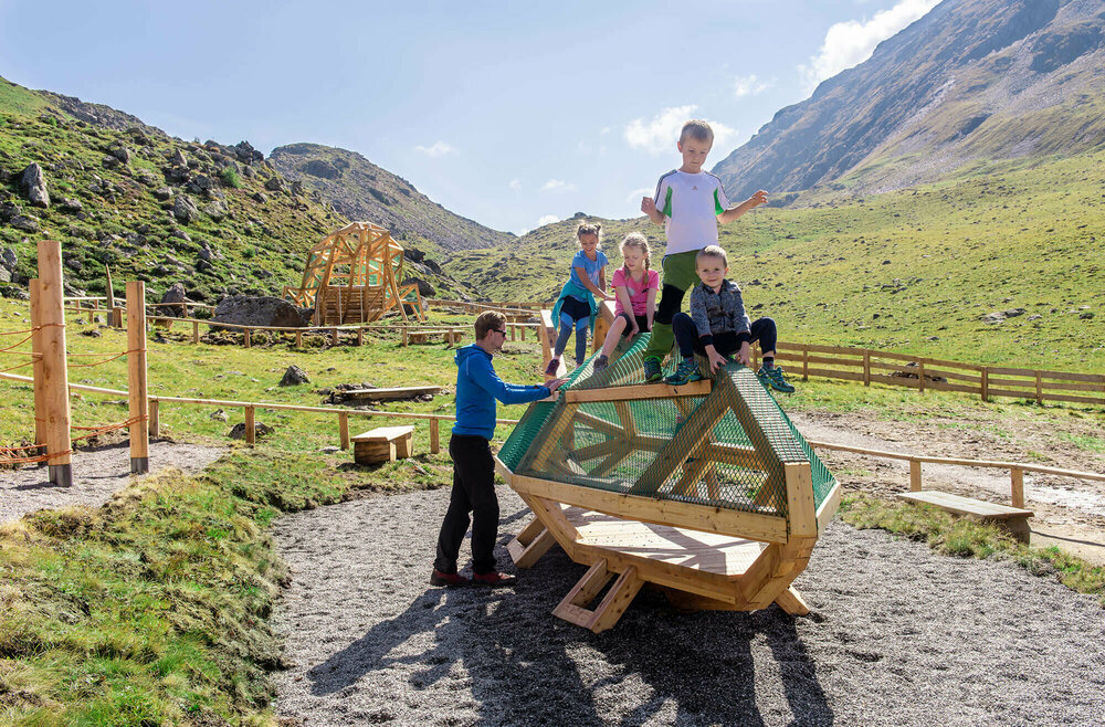 Spielende Kinder beim Wassermythos Ochsenlacke im Skizentrum St. Jakob i. D.. Sie klettern auf einem Spielgerät, was ihnen sichtlich viel Spaß bereitet. Die Sonne lässt die Umgebung und die dahinterliegende Bergwelt in einem warmen Licht erstrahlen.