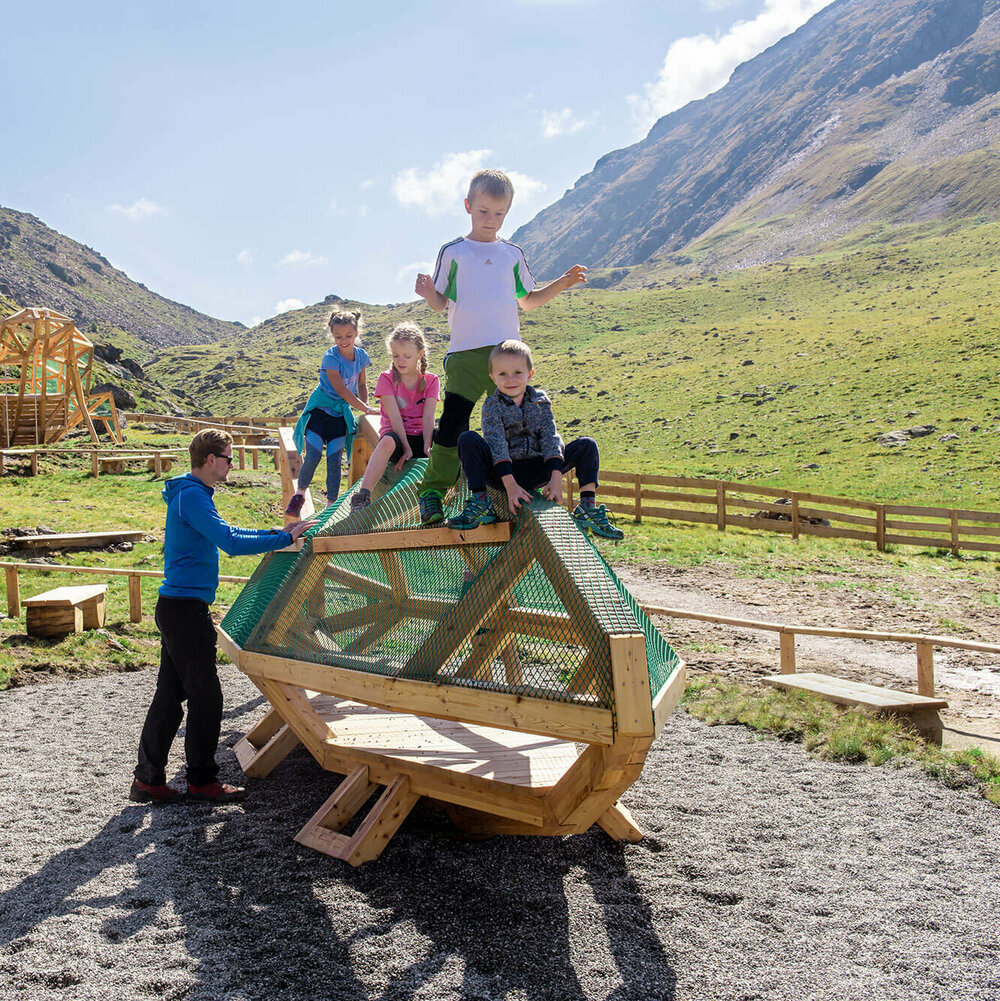 Wassermythos Ochsenlacke St. Jakob Brunnalm Spielende Kinder beim Wassermythos Ochsenlacke im Skizentrum St. Jakob i. D.. Sie klettern auf einem Spielgerät, was ihnen sichtlich viel Spaß bereitet. Die Sonne lässt die Umgebung und die dahinterliegende Bergwelt in einem warmen Licht erstrahlen.