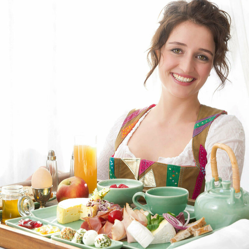 Frühstück Gutschein Dame in Dirndl serviert auf einem großen Tableau ein reichhaltiges Osttirol Frühstück mit Speck, Schinken, Käse, Ei, verschiedene Marmeladen und Honig und noch vieles Regionales mehr.