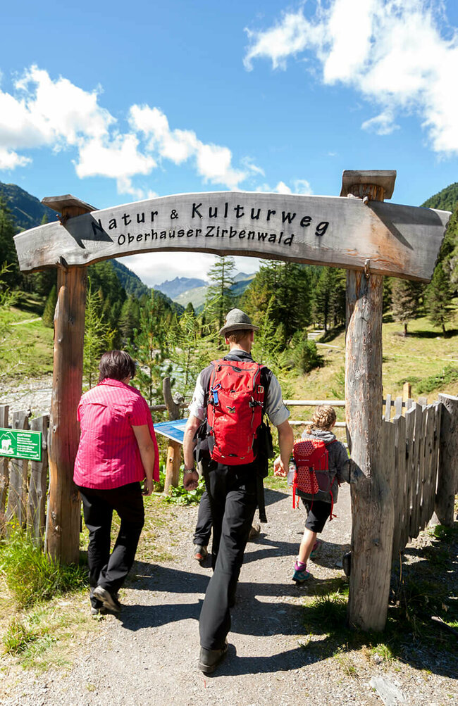 Oberhauser Zirbenwald Eine Familie in Wanderkleidung geht durch das aus Holz gebaute Eingangstor am Oberhauser Zirbenwald.